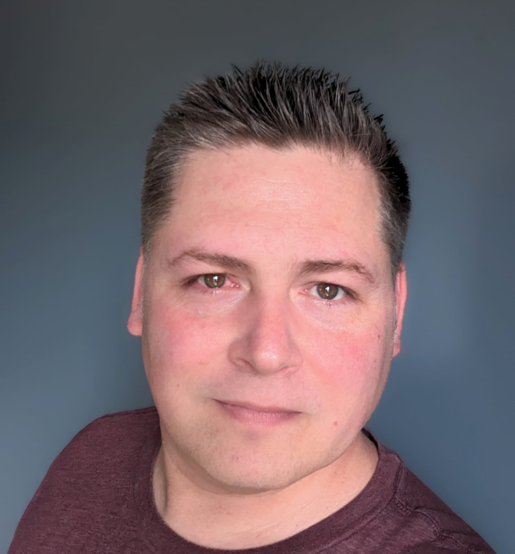 The image shows a man with short, neatly styled dark hair. He has fair skin and is wearing a maroon shirt. He is looking directly at the camera with a neutral expression and a slight smile. The background is a solid, muted gray-blue color, keeping the focus on his face.