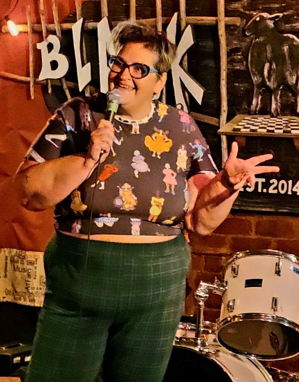 The image shows a woman performing stand-up comedy on a stage. She has short hair and is wearing glasses. Her outfit includes a colorful patterned top with various cartoon characters and high-waisted green plaid pants. She is holding a microphone with one hand and making a peace sign with the other, smiling and engaging with the audience. The background features a brick wall with string lights and musical instruments, including a drum set, creating a cozy and lively atmosphere.