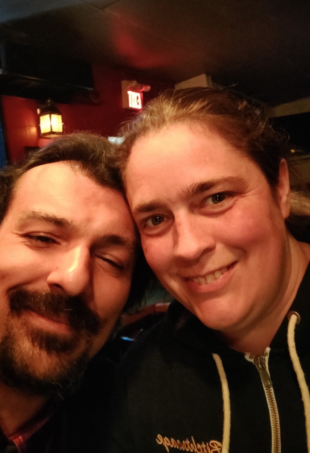 The image is a close-up selfie of a man and a woman. They are smiling and appear to be in a cozy, dimly lit indoor setting. The man has short dark hair and a beard, and his eyes are partially closed as he smiles. The woman has light brown hair pulled back and is wearing a hoodie. The background includes warm lighting and an exit sign, adding to the intimate, casual atmosphere.