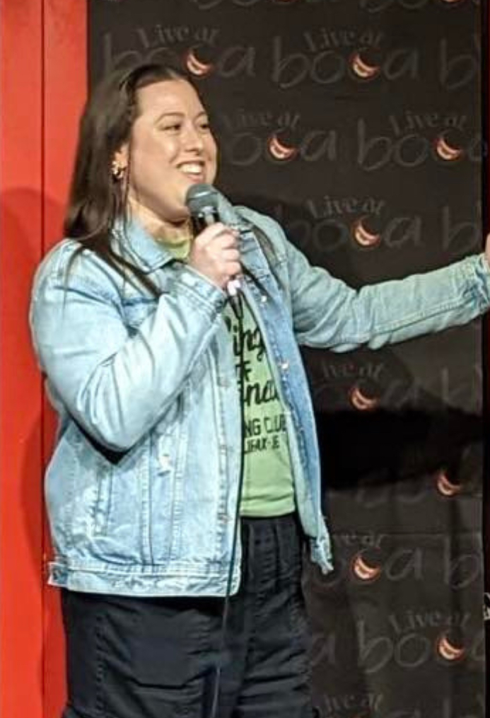 The image shows a woman performing stand-up comedy on stage. She has long dark hair and is wearing a light denim jacket over a green shirt, black cargo pants, and white sneakers. She is holding a microphone with one hand and the mic stand with the other, smiling as she engages with the audience. The backdrop features the logo "Live at Boa" and some lighting, creating a cozy comedy club atmosphere. The stage area has a red door and wooden floor, adding warmth to the setting.