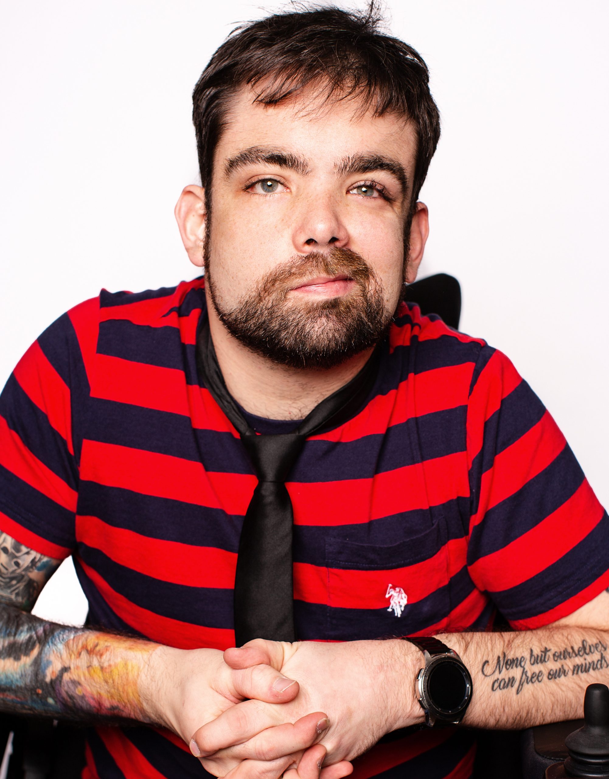 The image shows a man with short dark hair and a beard, wearing a red and navy striped shirt, a black necktie, and jeans. He is sitting and has his hands clasped in front of him. He has tattoos on his arms, one of which has text that reads, "None but ourselves can free our minds." He is looking directly at the camera with a calm, thoughtful expression. The background is plain and white, keeping the focus on the man.