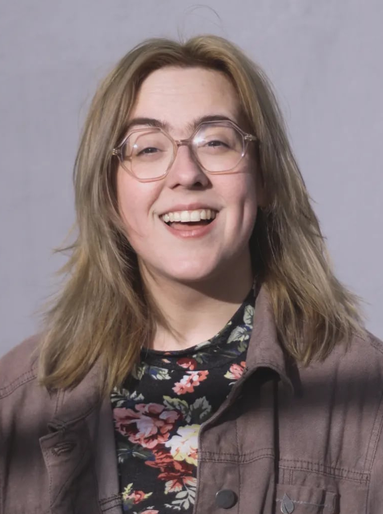 The image shows a smiling woman with light brown hair that falls just below her shoulders. She is wearing glasses and a floral-patterned top underneath a brown jacket. The background is plain and gray, focusing attention on her happy and approachable expression.