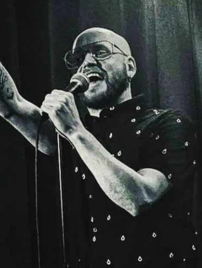 The image shows a man performing stand-up comedy on stage in a black and white setting. He is bald and has a beard, wearing glasses and a patterned short-sleeved shirt. He is holding a microphone with one hand and pointing upwards with the other, engaging energetically with the audience. The background is dark, enhancing the focus on the performer. His stance and expression suggest he is in the middle of delivering a punchline or an animated part of his act.