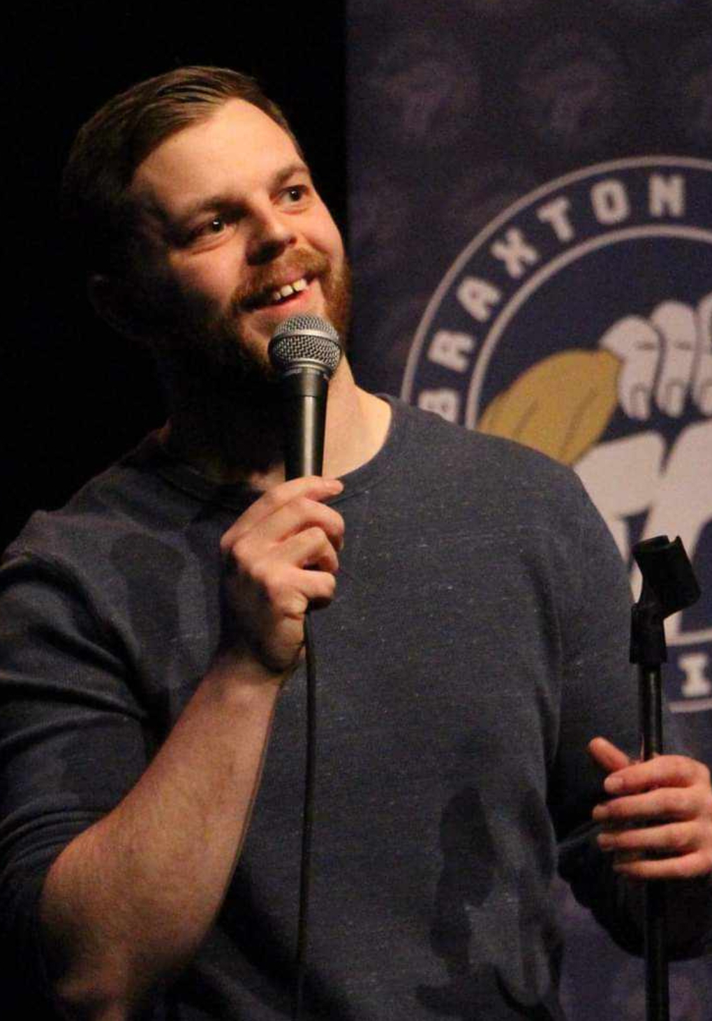 The image shows a man performing stand-up comedy on stage. He has short hair and a beard and is wearing a dark long-sleeve shirt. He is holding a microphone and smiling, looking engaged with the audience. Behind him is a banner with the logo of the "Braxton Comedy Festival," featuring an illustration of a fist holding a yellow chicken. The background is dark, highlighting the performer and the banner. The overall atmosphere suggests he is in the middle of a lively and enjoyable performance