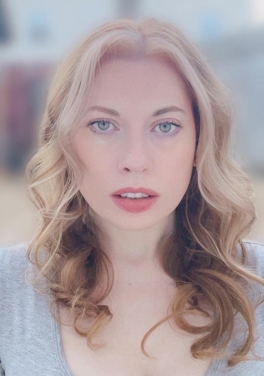 The image shows a woman with long, wavy blonde hair. She is wearing a gray short-sleeved top and has a neutral expression on her face. Her eyes are light-colored, and she has a calm and focused gaze. The background is blurred, featuring outdoor elements such as a fence and buildings, suggesting she is outside. The overall atmosphere is serene and natural.