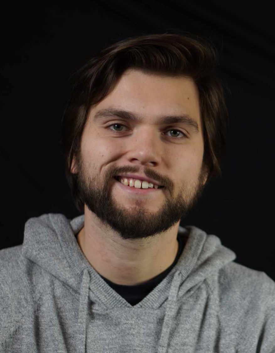 The image shows a man with short dark hair and a beard, smiling at the camera. He is wearing a light gray hoodie. The background is plain black, keeping the focus on his face. His expression is friendly and approachable.