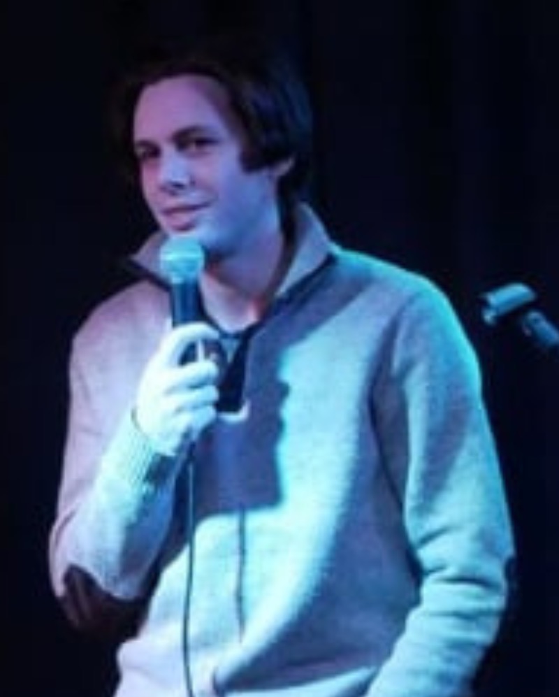 The image shows a man performing stand-up comedy on stage. He has medium-length dark hair and is wearing a light-colored sweater and dark pants. He is holding a microphone with one hand and has the other hand in his pocket. The background is dark, and he is illuminated by stage lighting, creating a focused and intimate atmosphere. He has a relaxed and confident expression, suggesting he is engaging comfortably with the audience.