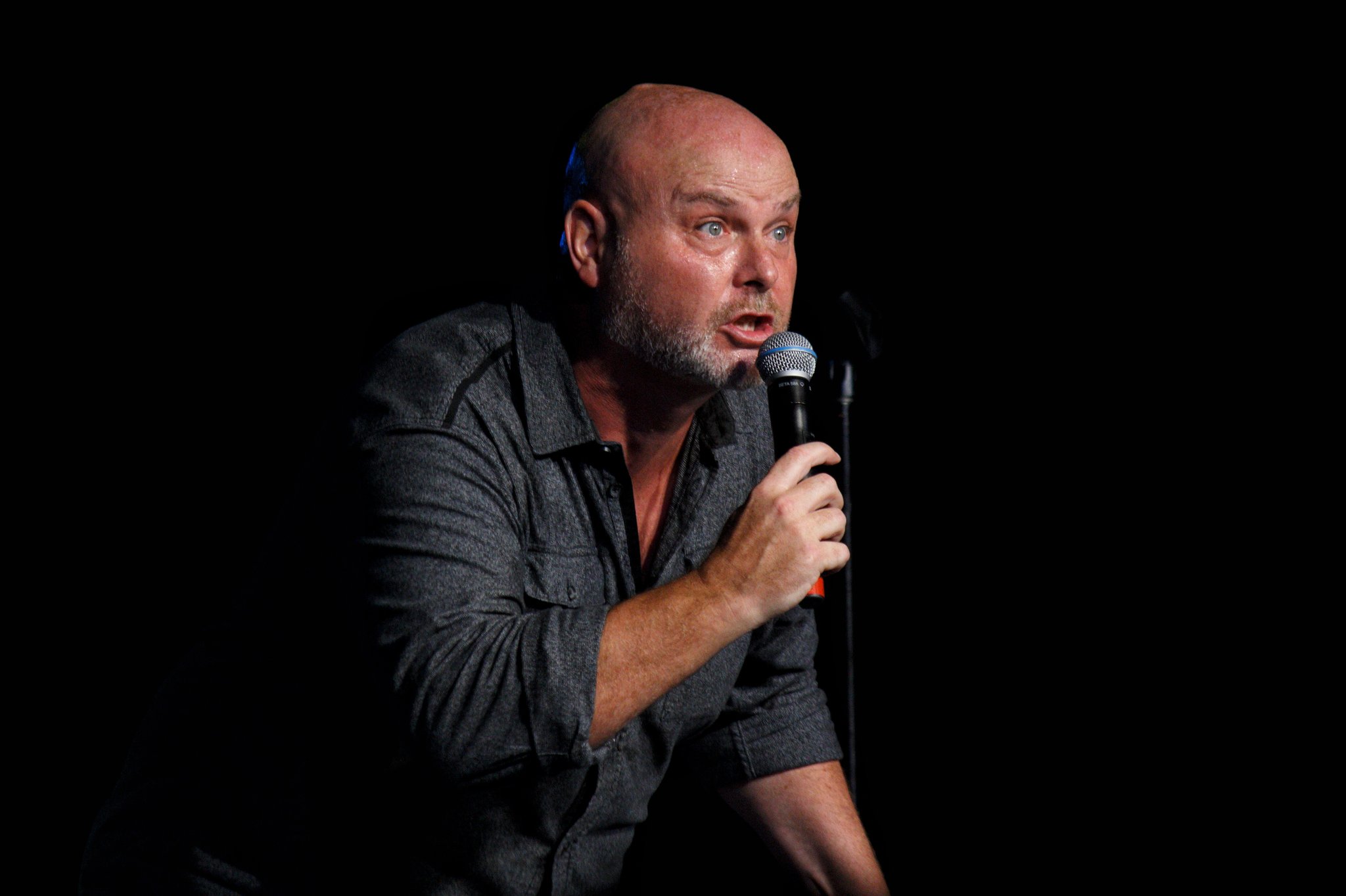 The image shows a bald man with a beard performing stand-up comedy on stage. He is holding a microphone in one hand and appears to be in the middle of delivering a joke or story, with an animated expression on his face. He is wearing a dark, long-sleeve shirt, and the background is completely black, highlighting him as the focal point of the image. The lighting emphasizes his features and creates a dramatic, engaging atmosphere typical of a comedy performance.