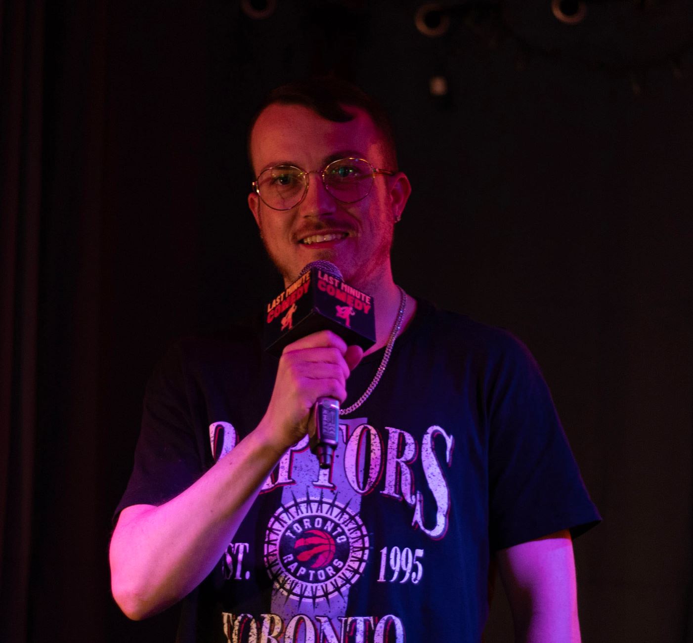 Subject Person: A man with glasses and short hair. Appearance Clothing: He is wearing a black T-shirt with "Toronto Raptors" and the Raptors logo printed on it. Facial Features: He has short hair and is wearing round glasses. Expression: He is smiling. Accessories: He is holding a microphone with a "Last Minute Comedy" label on it, suggesting he is performing or speaking. Background Lighting: The background is dark, and the lighting highlights the person. Additional Elements: There are no additional elements visible in the background, keeping the focus on the person. This image captures a moment of a man enjoying his time on stage, with the microphone and lighting suggesting a performance or comedic act.