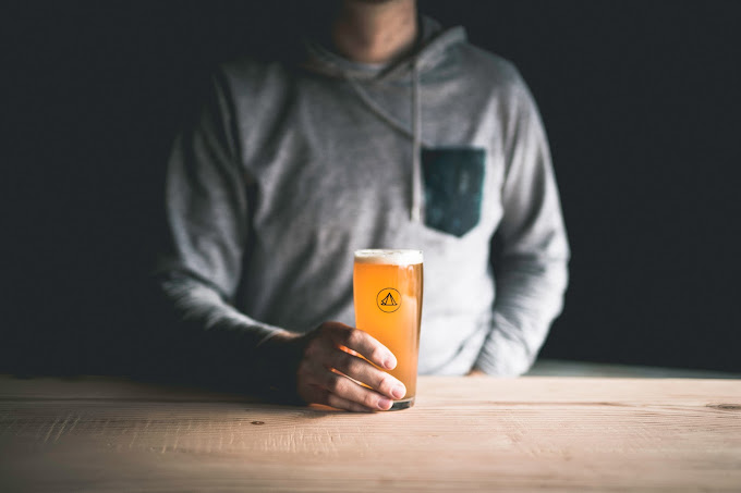 The image shows a person wearing a gray hoodie with a dark pocket, holding a pint of beer. The beer is a light amber color and is slightly cloudy, likely indicating a craft or unfiltered variety. The glass has a small logo on it, and the person is seated at a wooden table. The background is dark, creating a focus on the person and the beer, giving the scene a cozy, intimate feel.