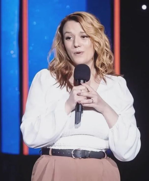 The image shows a woman with shoulder-length, wavy blonde hair performing stand-up comedy. She is holding a microphone with both hands and has a friendly, engaging expression. She is wearing a white blouse with long sleeves and a pair of high-waisted, light brown pants with a black belt. The background features vertical colored lights, creating a vibrant and lively stage atmosphere.