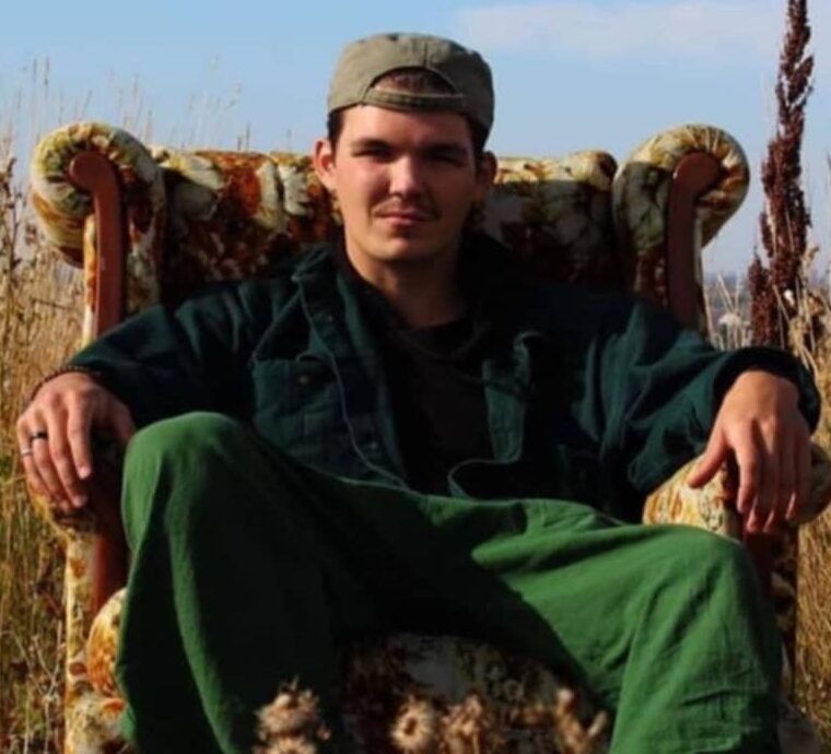 The image shows a young man sitting in an old-fashioned, upholstered armchair placed outdoors in a field. He is wearing a green jacket, matching green pants, and a backwards baseball cap. His expression is relaxed and confident as he leans back in the chair with his hands resting on the armrests. The setting features tall grasses and wildflowers, creating a natural, serene atmosphere under a clear blue sky.