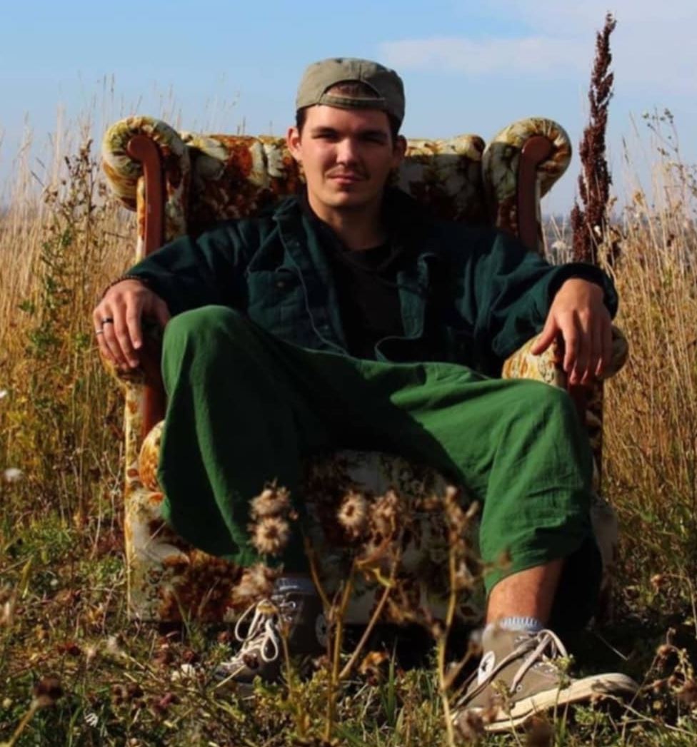 The image shows a young man sitting in an old-fashioned, upholstered armchair placed outdoors in a field. He is wearing a green jacket, matching green pants, and a backwards baseball cap. His expression is relaxed and confident as he leans back in the chair with his hands resting on the armrests. The setting features tall grasses and wildflowers, creating a natural, serene atmosphere under a clear blue sky.