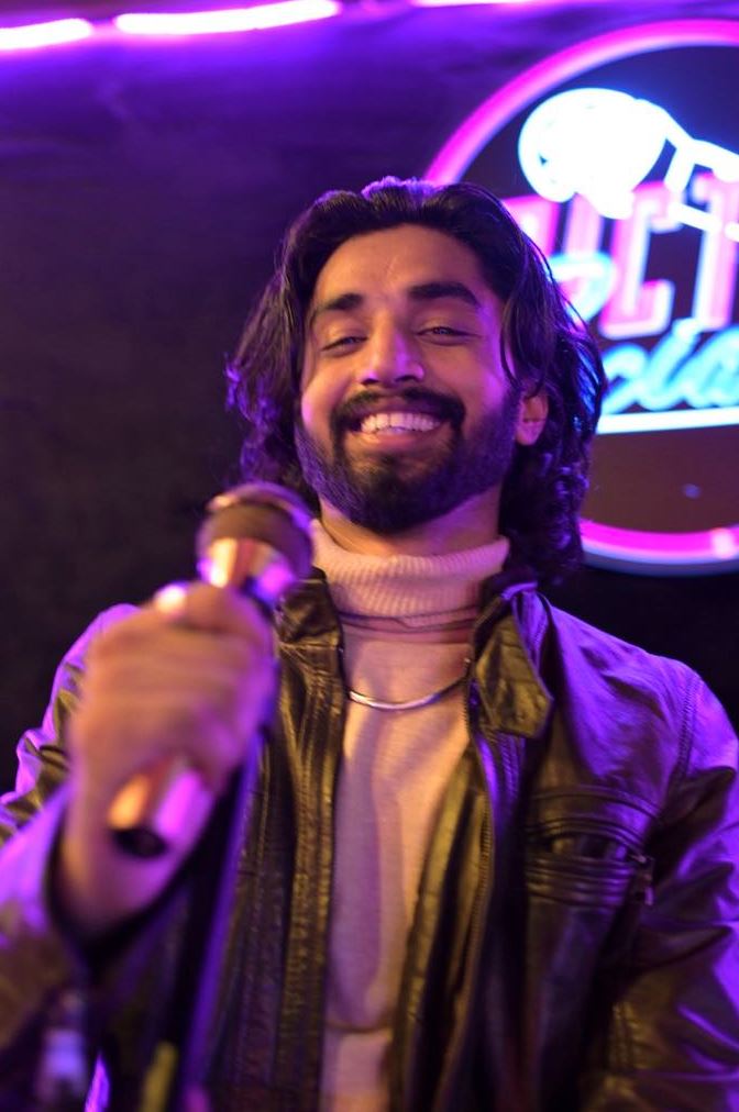 Subject Person: A man with long hair and a beard. Appearance Clothing: He is wearing a light-colored turtleneck and a leather jacket. Facial Features: He has long, dark hair and a beard. Expression: He is smiling warmly. Accessories: He is holding a microphone, suggesting he is performing or speaking. Background Lighting: The background is dark with purple lighting, creating a vibrant and dynamic atmosphere. Additional Elements: There is a neon sign in the background, adding to the lively setting. This image captures a moment of a man enjoying his time on stage, with the colorful lighting and neon sign enhancing the energetic ambiance.