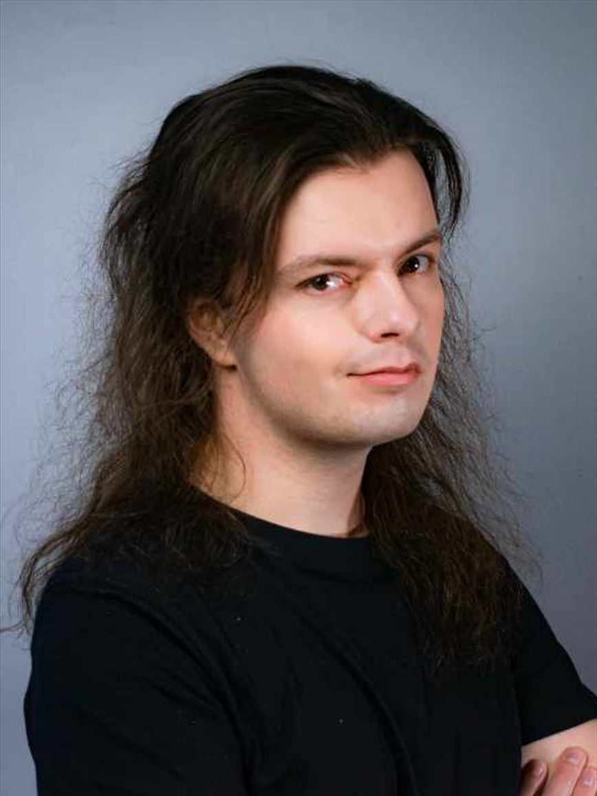 The image shows a man with long, dark, slightly wavy hair and fair skin. He is wearing a black shirt and has a subtle smile on his face. He is looking directly at the camera with a calm and composed expression. The background is plain and gray, keeping the focus on his face and upper body.