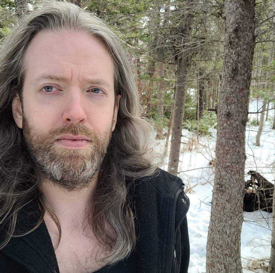 Subject Person: The person in the image appears to be a male with long hair and a beard. Appearance Hair: Long, grayish hair. Clothing: Wearing a dark jacket or coat. Expression: The expression is neutral, possibly slightly contemplative, with direct eye contact with the camera. Background Setting: The background suggests an outdoor environment, possibly a forest or wooded area. There is snow on the ground and trees in the background. This image captures a serene and natural moment, with the subject standing outdoors in a wintery landscape, providing a calm and thoughtful atmosphere.