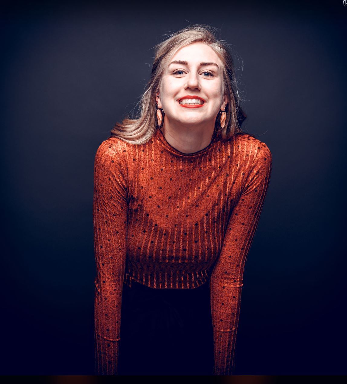 Subject Person: A woman with blonde hair and a bright smile. Appearance Clothing: She is wearing a shiny, long-sleeved, rust-colored top with a dotted pattern. Facial Features: She has long blonde hair, styled to one side, and is wearing large earrings. Expression: She has a wide, genuine smile. Pose: She is slightly leaning forward, adding a dynamic feel to the image. Background Lighting: The background is dark, which contrasts with her brightly colored top and highlights her facial features. Additional Elements: There are no additional elements in the background, ensuring the focus remains on the subject. This image captures a moment of joy and confidence, with the woman's bright smile and vibrant outfit standing out against the dark background.