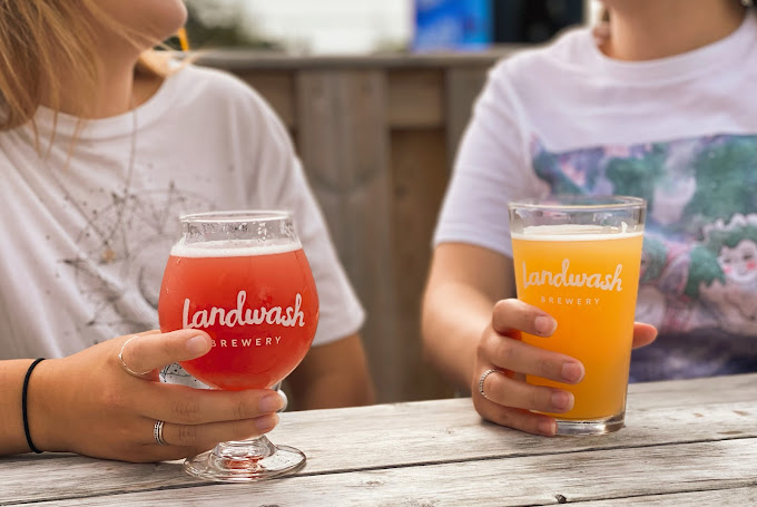 The image shows two people sitting at a wooden table, each holding a glass of beer. The glass on the left contains a red beer, while the glass on the right contains a hazy yellow beer. Both glasses have the "Landwash Brewery" logo printed on them. The people are wearing casual white T-shirts, and the setting appears to be outdoors, suggesting a relaxed and enjoyable atmosphere, possibly at a brewery or a casual gathering.