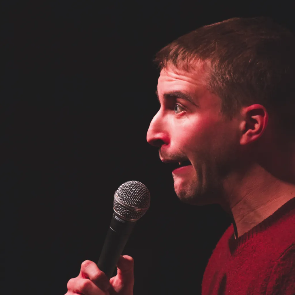 The image shows a man with short dark hair performing stand-up comedy on stage. He is holding a microphone close to his mouth with both hands and has an expressive, animated facial expression, suggesting he is in the middle of delivering a joke or a punchline. He is wearing a dark-colored top. The background is dark, focusing attention on the performer and his expressive demeanor.