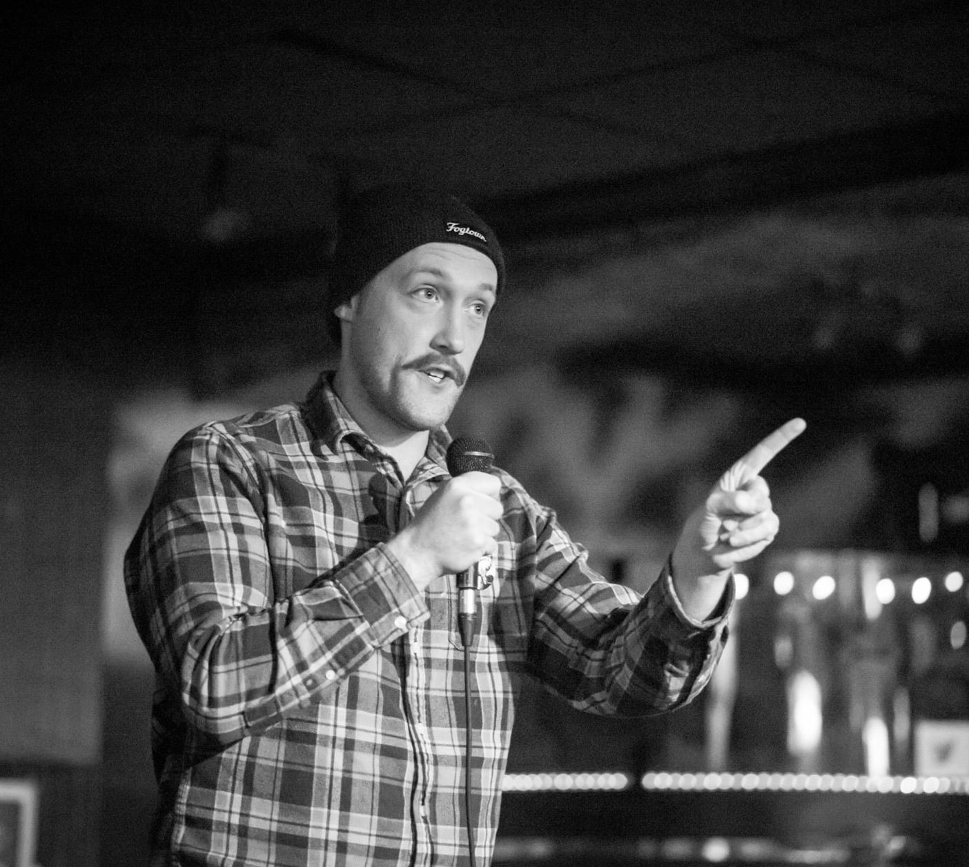 Subject Person: A man performing stand-up comedy. Appearance Clothing: He is wearing a plaid shirt and a beanie hat. Facial Expression: He has a focused and engaged expression, interacting with the audience. Accessories: He is holding a microphone in one hand. Environment Background: The background appears to be a dimly lit bar or club setting, creating an intimate atmosphere. Stage Setup: There are lights in the background, possibly from the bar or stage setup, creating a warm and inviting ambiance. Lighting: The lighting is focused on the performer, highlighting his presence on the stage.