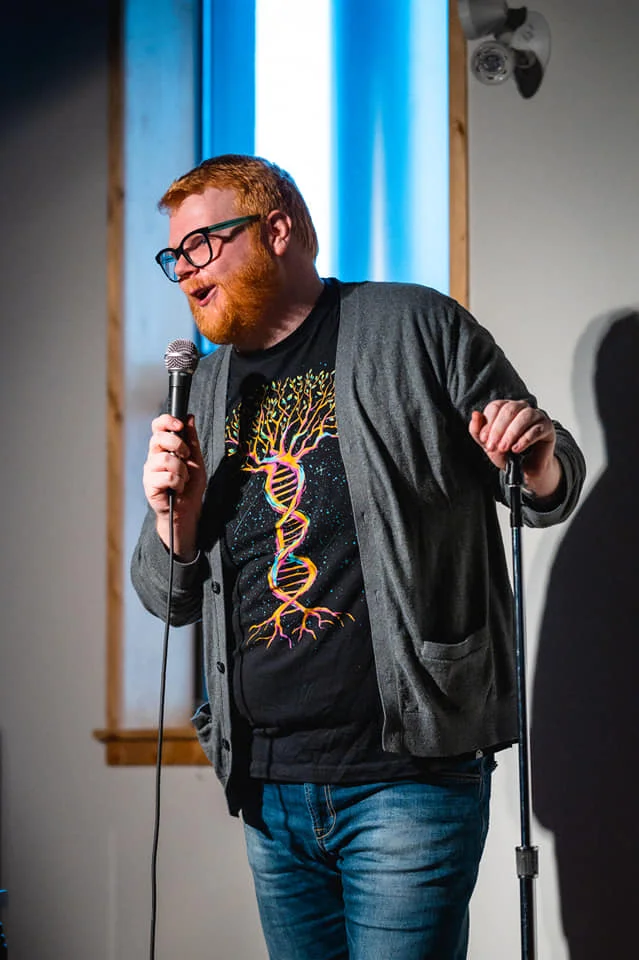 The image shows a man with red hair and a beard performing stand-up comedy. He is wearing glasses, a gray cardigan, and a black T-shirt with a colorful DNA double helix design that transitions into tree roots. He is holding a microphone in one hand and the stand in the other, smiling and speaking, suggesting he is in the middle of a joke or story. The background features a lit window, creating a bright and engaging setting. The overall atmosphere is lively and engaging, characteristic of a comedy performance.