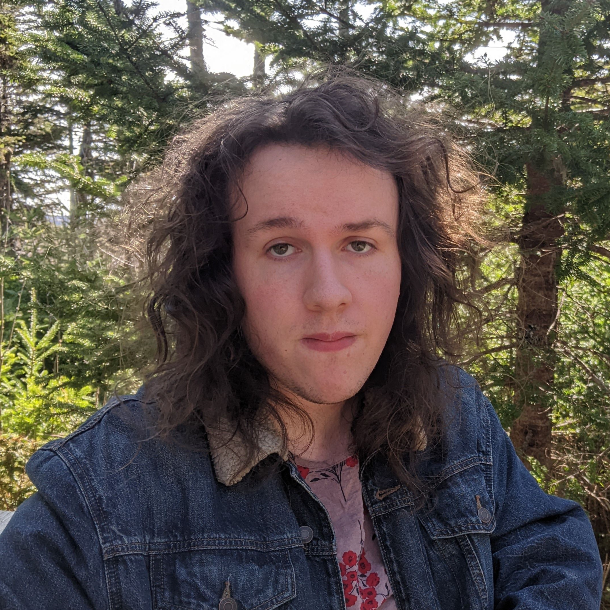 The image shows a young person with long, wavy dark hair. They are wearing a denim jacket with a shearling collar over a floral-patterned top. Their expression is calm and relaxed, looking directly at the camera. The background features lush green trees, suggesting the photo was taken outdoors in a forest or park setting. The overall atmosphere is serene and natural.