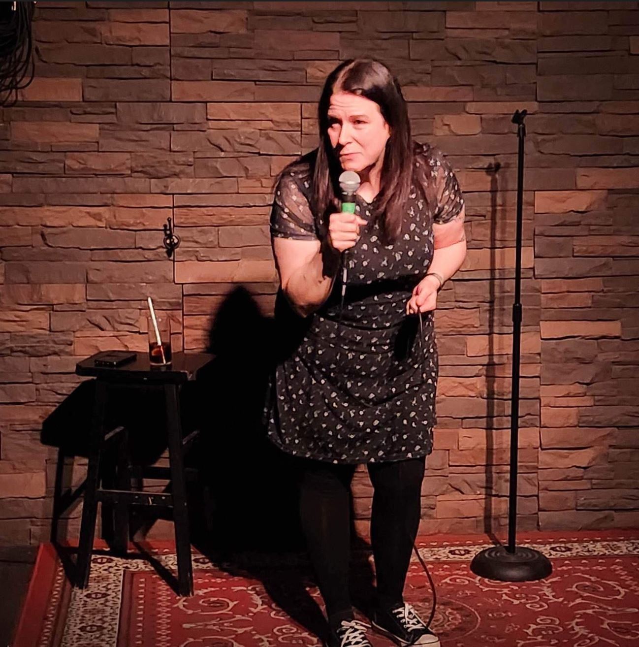 Subject Person: A woman performing stand-up comedy. Appearance Clothing: She is wearing a dark dress with a floral pattern, black tights, and black sneakers. Facial Expression: She has a focused and animated expression, engaging with the audience. Accessories: She is holding a microphone in one hand. Environment Background: The backdrop consists of a brick or stone wall, giving a rustic and intimate vibe to the setting. Stage Setup: There is a small table or stool next to her with a drink and some items on it. A microphone stand is positioned behind her. The stage has a red patterned carpet. Lighting: The stage lighting is focused on her, highlighting her presence and creating a shadow against the wall. This image captures a moment of live comedy, showcasing the performer in an intimate and cozy setting, with an emphasis on her engaging stage presence.