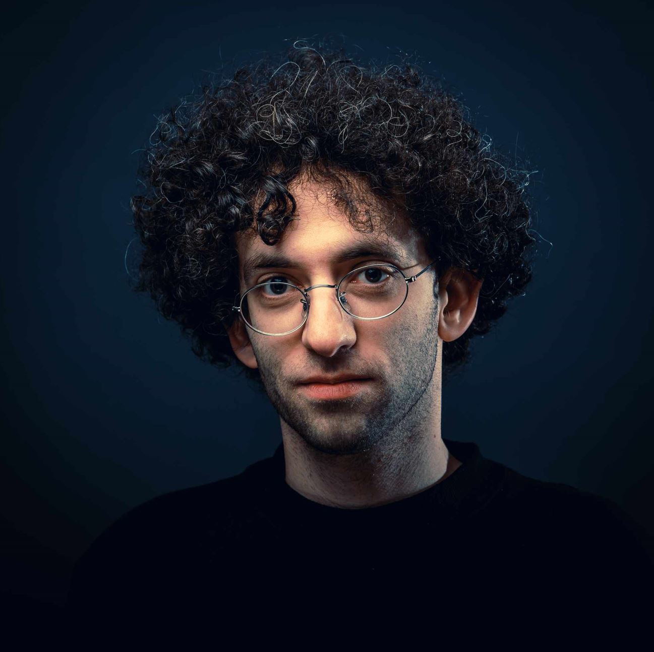 Subject Person: A man with curly hair and glasses. Appearance Clothing: He is wearing a dark top. Facial Features: He has distinct curly hair and is wearing round glasses. Expression: His expression appears calm and neutral. Background Lighting: The background is dark, which creates a contrast that highlights the subject's face and hair. This image captures a close-up portrait of the man, emphasizing his facial features and the texture of his curly hair. The dark background and lighting focus the viewer's attention on the subject.