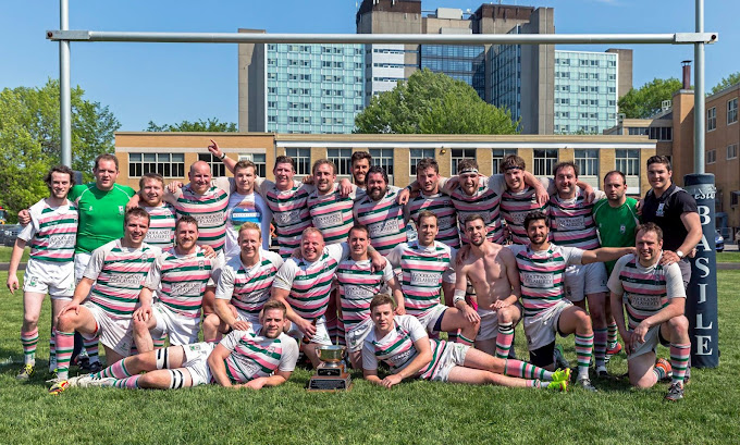 The image shows a group of rugby players posing together on a field. They are all wearing matching striped jerseys in white, green, pink, and black, with white shorts and rugby socks. The team members are smiling and appear to be celebrating a victory, with one player holding a trophy at the front. The group is positioned under a rugby goal post, and behind them is a building with multiple windows, set against a backdrop of green trees and a clear sky. The overall atmosphere is joyful and triumphant, indicating a successful match or tournament win.