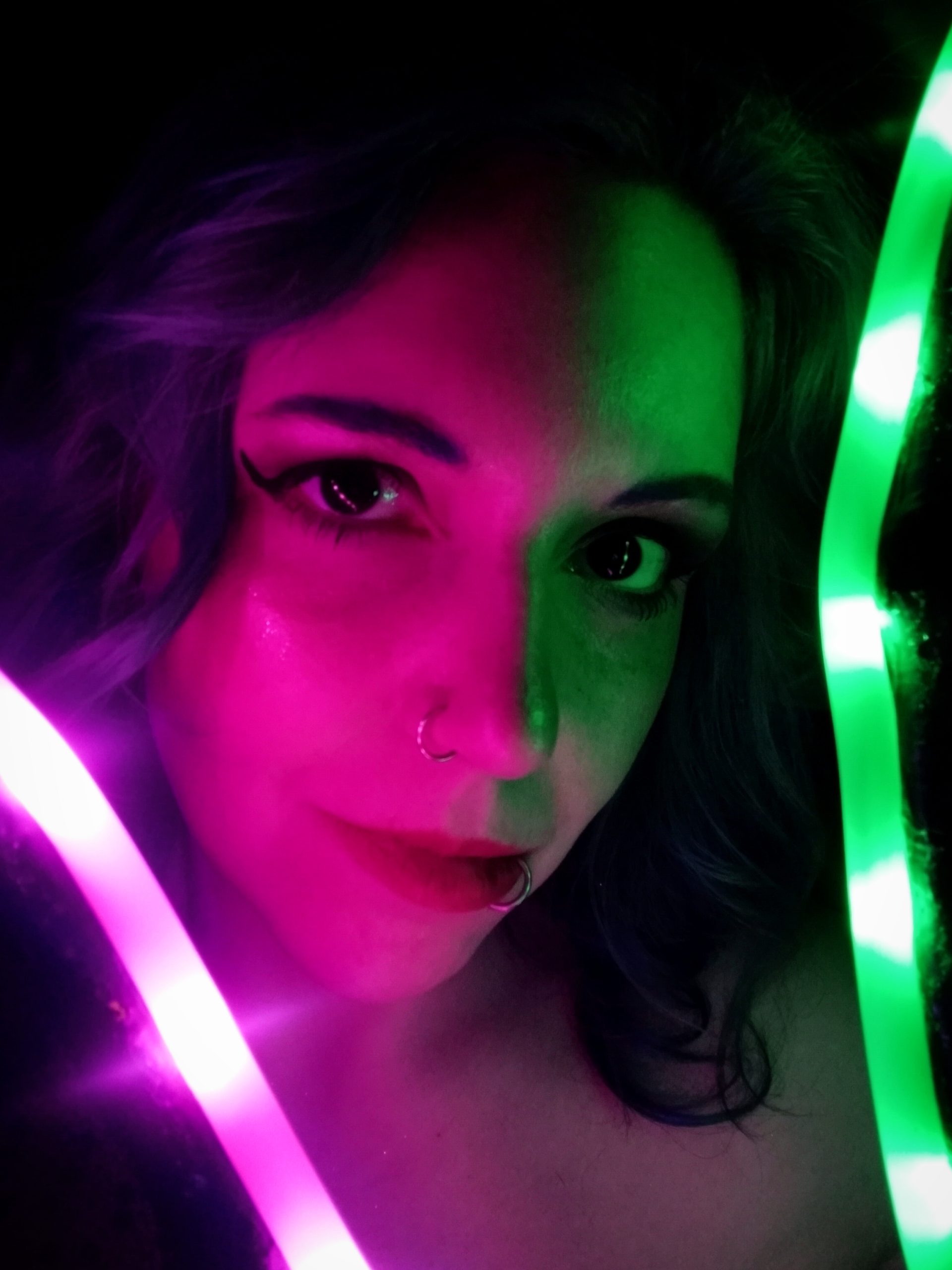 Subject Person: A woman with distinctive makeup and piercings. Appearance Facial Features: She has bold eyeliner, a nose ring, and a lip ring. Lighting: The image is creatively lit with neon lights, casting green and pink hues across her face. Expression: She has a calm and confident expression, looking directly at the camera. Background Lighting: The background is dark, making the neon lights and her features stand out more dramatically. Additional Elements: There are bright neon lights, one pink and one green, framing her face and adding to the artistic effect of the photograph. This image captures a striking and artistic portrait, with the neon lights providing a unique and vibrant contrast to the subject's features.