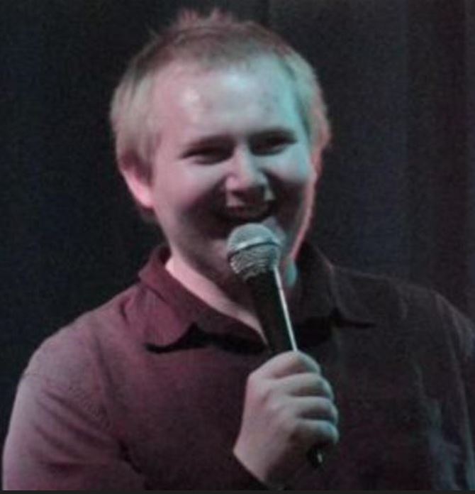 Subject Person: The person in the image appears to be a male comedian, smiling and holding a microphone, suggesting he is mid-performance. Appearance Hair: Light-colored, short hair. Clothing: Wearing a dark-colored shirt. Expression: Smiling, with a joyful and engaging expression, indicating he is likely making the audience laugh. Background Lighting: The lighting has a soft, possibly stage-like effect, with a slightly dim background which helps to focus on the comedian. This image captures a candid moment of the comedian enjoying his time on stage, engaging with the audience through his performance.