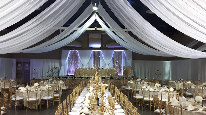 The image depicts a beautifully decorated banquet hall set up for an event. The ceiling is adorned with elegant white drapery, creating a grand and airy atmosphere. Long tables are arranged in rows, covered with white tablecloths and decorated with gold accents. Each table has gold chairs and neatly folded white napkins. The head table is positioned at the front, against a backdrop of sparkling gold and white fabric with soft lighting, enhancing the festive ambiance. The overall decor suggests a formal and luxurious event, such as a wedding reception or a gala.