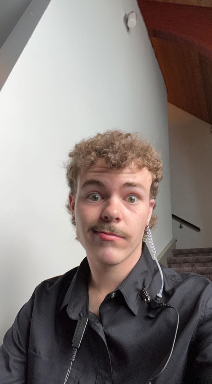 The image shows a young man with curly blond hair and a mustache, making a playful expression. He is wearing a black shirt and has an earpiece with a coiled cord attached to it, likely indicating he is working in some capacity where communication is needed. He is indoors, near a staircase, and the background features plain white walls and wooden accents. His expression and the setting suggest a casual, light-hearted moment.