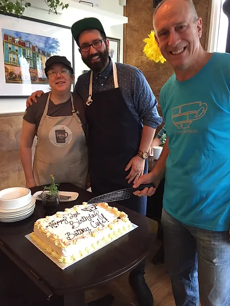 The image shows three people standing together and smiling in a celebratory setting at Battery Cafe. The person on the left is wearing a gray apron and a black cap. The person in the middle is wearing a blue shirt, a black apron, and a green cap, and has a beard and glasses. The person on the right is wearing a teal T-shirt with a coffee cup logo and is holding a knife. They are standing behind a table with a large rectangular cake that says "Happy Birthday Battery Cafe!" in black icing. There are plates and cups on the table, and the atmosphere looks festive and cheerful.