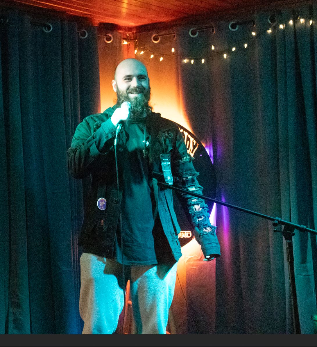 Subject Person: A bearded man performing stand-up comedy. Appearance Clothing: He is wearing a dark jacket with some distressed details on the sleeves and light-colored pants. Setting: The stage has dark curtains as the backdrop with string lights hanging above. There is also a microphone stand and a wooden chair visible to the side. Background Lighting: The stage is illuminated with a combination of blue and warm lights, creating a cozy atmosphere. The background includes a circular logo partially visible behind the performer. This image captures a moment from a comedy performance, emphasizing the relaxed and intimate setting of the venue.