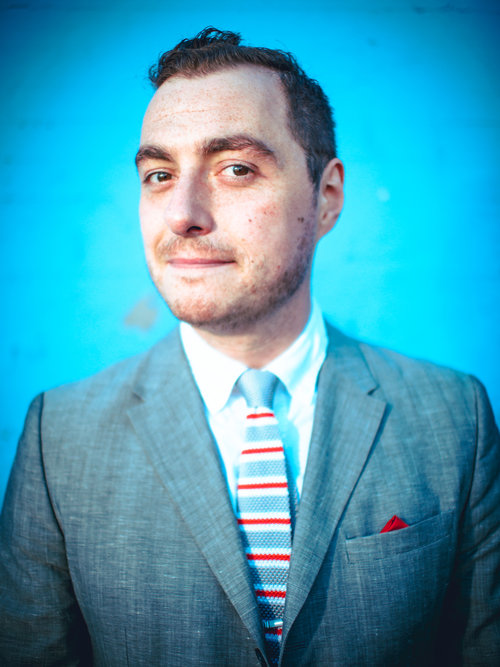 The image shows a man with short dark hair and a subtle smile. He is wearing a light gray suit, a white shirt, and a striped tie with colors including red and blue. He also has a red pocket square in his suit jacket pocket. The background is a bright blue wall, which contrasts with his attire and makes him stand out. His expression is confident and approachable.