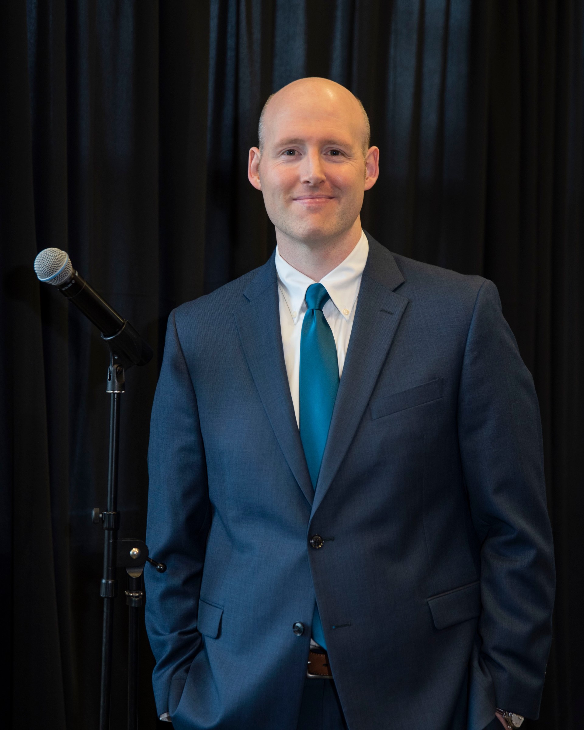 This image shows a man with a light complexion, who is bald and dressed in a formal suit. He is wearing a blue suit jacket, a white dress shirt, and a teal tie. He stands with one hand in his pocket and is smiling slightly, posing next to a microphone stand. The background features a black curtain, giving the setting a professional or event atmosphere.