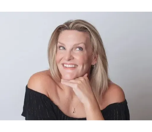 The image is a professional portrait of a woman named Fiona O'Brien. She has blonde hair, parted slightly off-center, and a friendly, engaging smile. Fiona's pose is relaxed and confident, with her chin resting on her right hand and her elbow on the table or surface. She is wearing an off-the-shoulder black top, revealing her shoulders and collarbones. The background is a plain, light color, which keeps the focus on her face and expression. Fiona's makeup is subtle and enhances her natural features, adding to the polished look of the portrait.