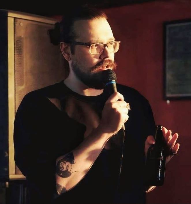 The photo features a male comedian, Kevin Coffey, on stage. He has a distinctive mustache and beard, and he is wearing glasses. His hair is pulled back into a bun. He is holding a microphone in one hand and a bottle in the other. The lighting is warm, casting a glow on his face. He is dressed in a dark shirt, and his right arm has a visible tattoo. The background is dimly lit, focusing attention on him as he performs.