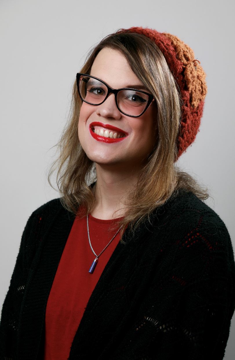 The image features a person with a cheerful expression, wearing a red top and a black cardigan. They have a beige and brown knitted hat with a decorative flower on the side. They have long, light brown hair and are wearing black-rimmed glasses. They also have a bright red lipstick and are smiling broadly. They are wearing a necklace with a blue pendant. The background is plain and light-colored, keeping the focus on the subject.