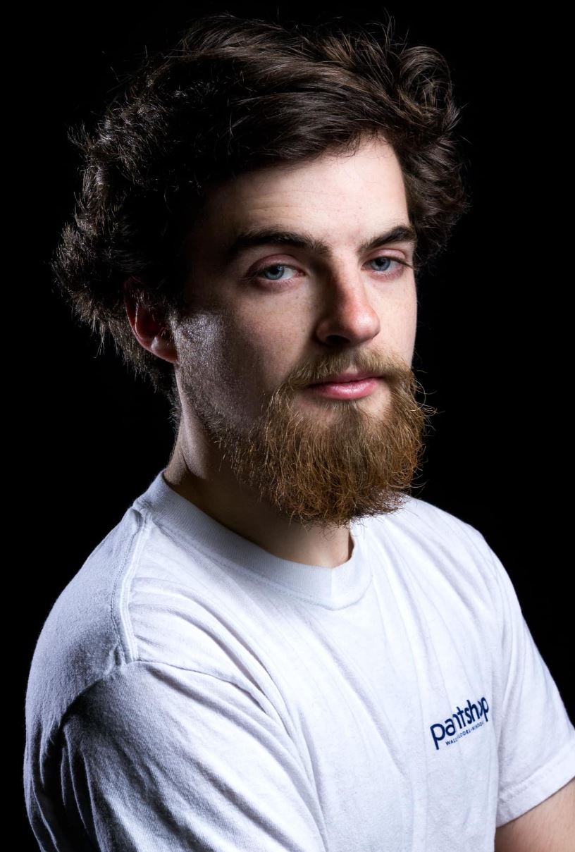 This image shows a young man with a light complexion, a bushy brown beard, and tousled brown hair. He is wearing a white T-shirt with a small logo on the left chest that reads "Paintshop." He has a serious expression and is looking directly at the camera. The background is black, providing a stark contrast to his light-colored clothing and skin.