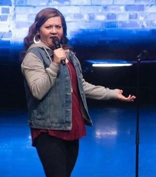 The image shows Lisa Baker performing on stage. She is holding a microphone in her right hand and gesturing with her left hand. Lisa is wearing a denim vest over a maroon top and dark pants. Her expression appears animated as she engages with the audience. The background features a blue-lit wall, possibly with a brick pattern, and stage lighting.