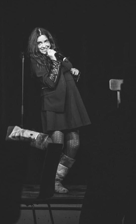 The black and white photo features a female comedian on stage. She has long, wavy hair and is smiling while holding a microphone close to her mouth. She is wearing a dark jacket over a dress, patterned tights, and tall boots. One of her legs is playfully lifted behind her as she leans slightly to the side, adding a lively and engaging touch to her performance. The background is dark, drawing attention to her expressive pose and joyful expression.