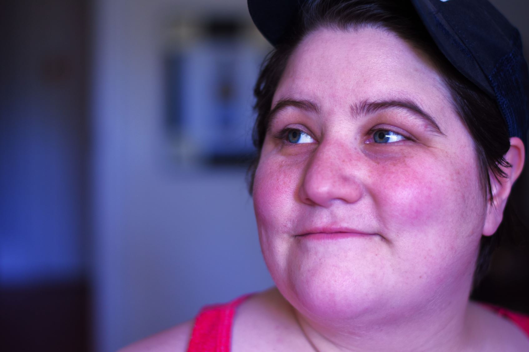 This image is a close-up portrait of a person with a light complexion. They have short, dark hair and are wearing a black baseball cap. The person is looking off to the right with a slight, serene smile. Their cheeks are slightly flushed, and they have a few freckles. They are dressed in a bright pink tank top. The background is softly blurred, putting the focus entirely on their face.