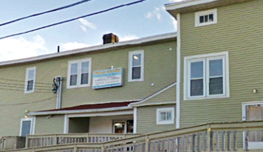 This image shows the exterior of a two-story building with light green siding and white window frames. There is a sign on the upper part of the building that likely displays the name of the establishment, though the details are hard to make out from this angle. The structure has a ramp or walkway leading up to an entrance, suggesting accessibility for those with mobility needs. The building has a homey, somewhat older architectural style, and power lines are visible across the top. The clear sky suggests a calm, pleasant day.