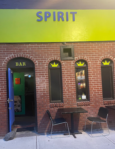 The image shows the exterior of a bar named "Spirit." The building has a brick facade with three arched windows, and a small sign above the entrance door that reads "BAR" in yellow letters. The door to the bar is open, revealing a glimpse of the interior. Above the windows and door, there is a bright yellow-green sign with the name "SPIRIT" written in bold blue letters. In front of the building, there is a small table with two chairs on either side, suggesting an outdoor seating area. The overall atmosphere appears inviting, with the open door and cozy setup outside.