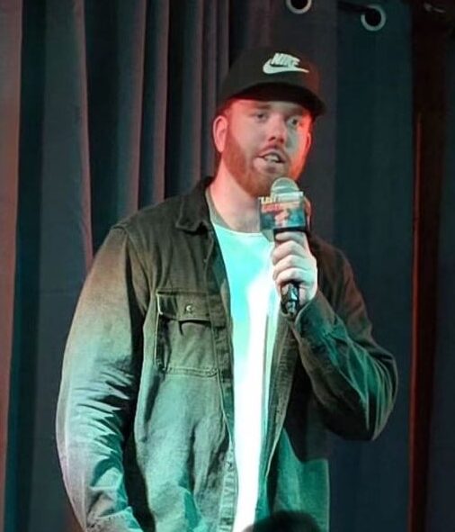 The image shows a man on stage holding a microphone, appearing to be mid-performance. He is wearing a black Nike cap, a dark denim jacket over a white t-shirt, and dark jeans. The setting seems to be a small, intimate venue with a dark curtain as the backdrop, creating a focused atmosphere on the performer. The man has a reddish beard and is speaking into the microphone, likely engaging with the audience.