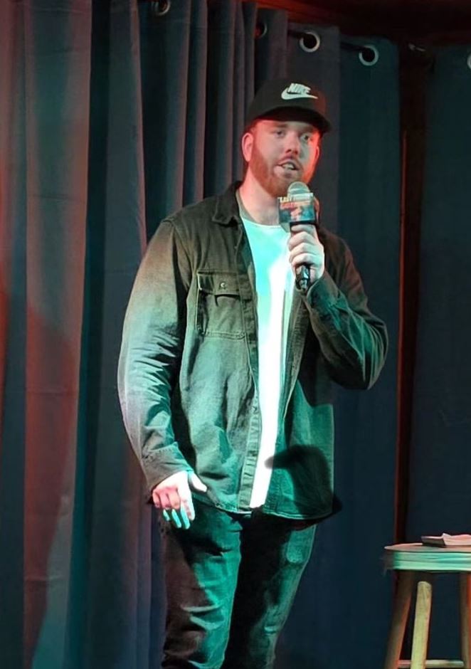 The image shows a man on stage holding a microphone, appearing to be mid-performance. He is wearing a black Nike cap, a dark denim jacket over a white t-shirt, and dark jeans. The setting seems to be a small, intimate venue with a dark curtain as the backdrop, creating a focused atmosphere on the performer. The man has a reddish beard and is speaking into the microphone, likely engaging with the audience.