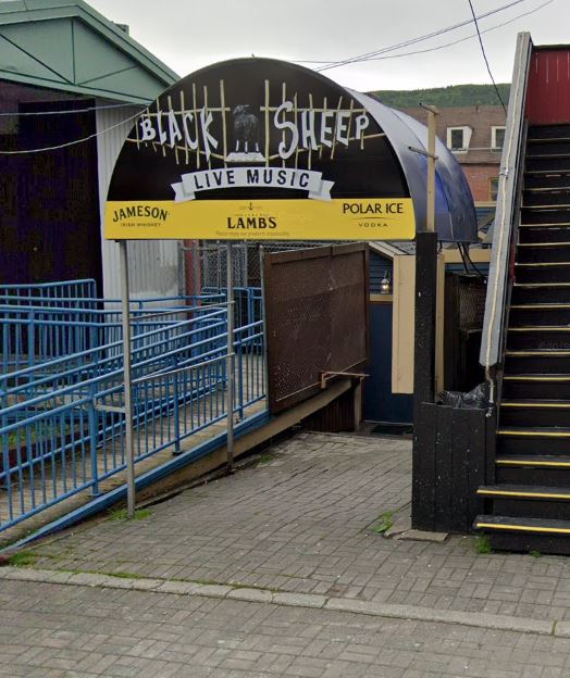 This image shows the entrance to The Black Sheep, a live music venue. The entrance is marked by an awning displaying the venue's name along with advertisements for Jameson, Lamb's, and Polar Ice vodka. The awning is arched and has a dark design, with "LIVE MUSIC" prominently displayed. The entrance appears to lead down a ramp or stairs, indicating it might be located below street level. A staircase to the right leads upward, possibly to another building or street level. The metal railings and signage suggest that this is a lively, casual venue, likely popular for music events, and potentially comedy shows as well.