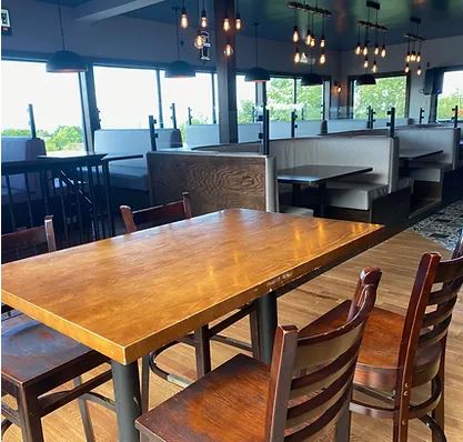 This photo showcases a cozy dining area with polished wooden tables and matching chairs. The space features booth seating along the windows, providing a comfortable and private dining experience with views of the surrounding greenery. Overhead lighting with an industrial design adds warmth and ambiance to the modern decor. This venue appears to be ideal for casual dining or hosting small events.